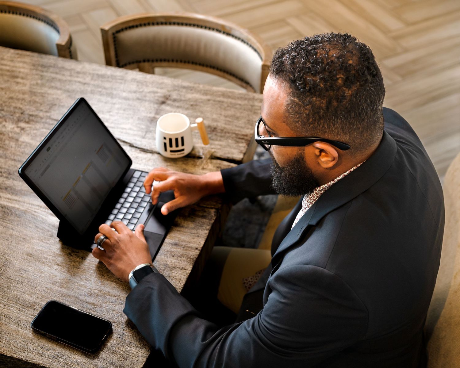 Business professional in dark suit working on digital tablet at wooden desk in office setting.