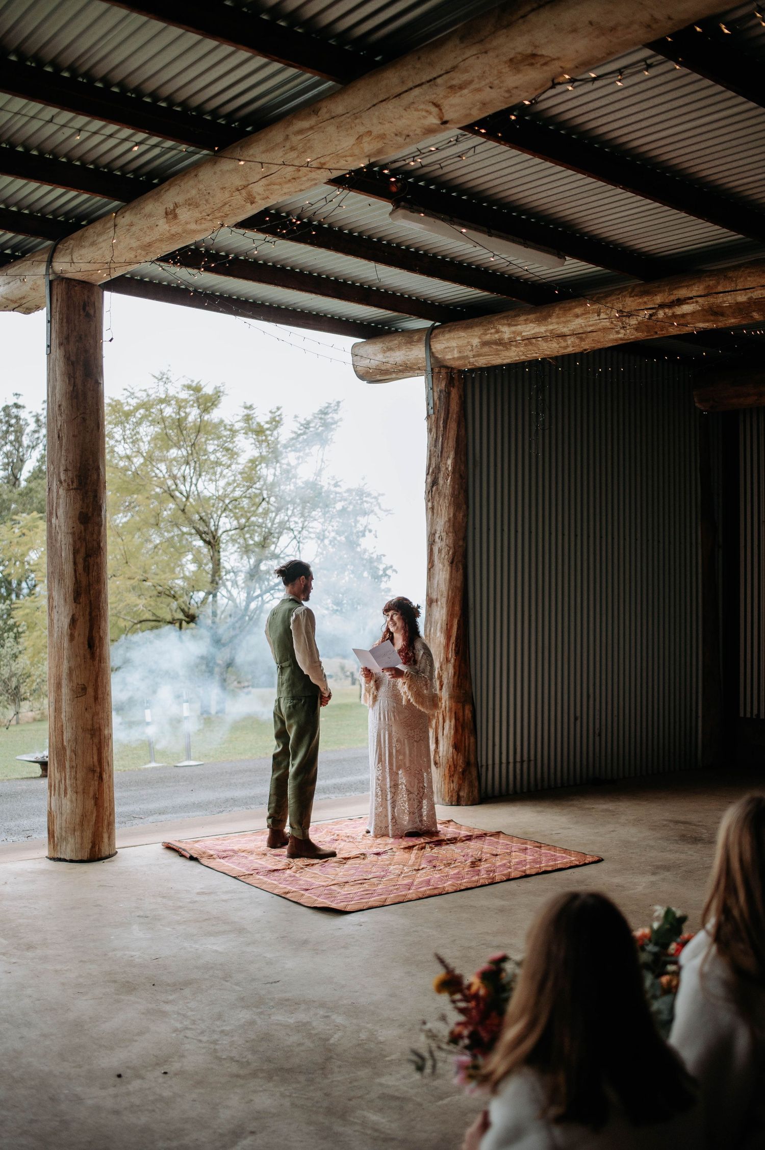 A couple stands together on a red rug under a rustic wooden porch during an intimate outdoor ceremony setting.