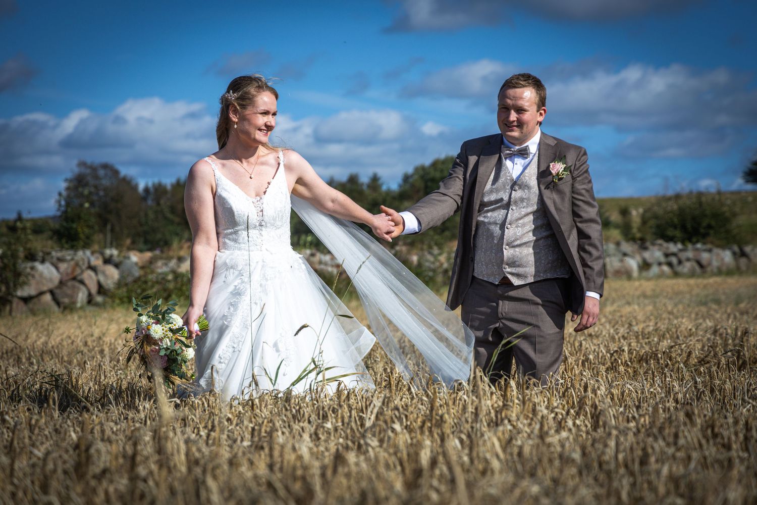 Bride and groom walk hand in hand through golden wheat field under bright blue sky.