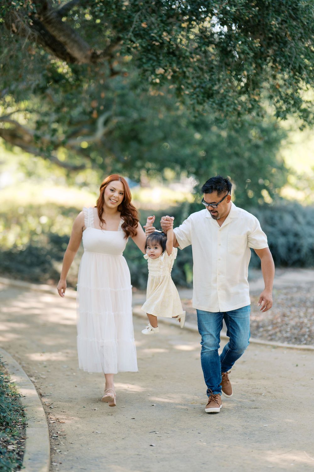 A family walks and plays together on a sunlit dirt path surrounded by green trees.