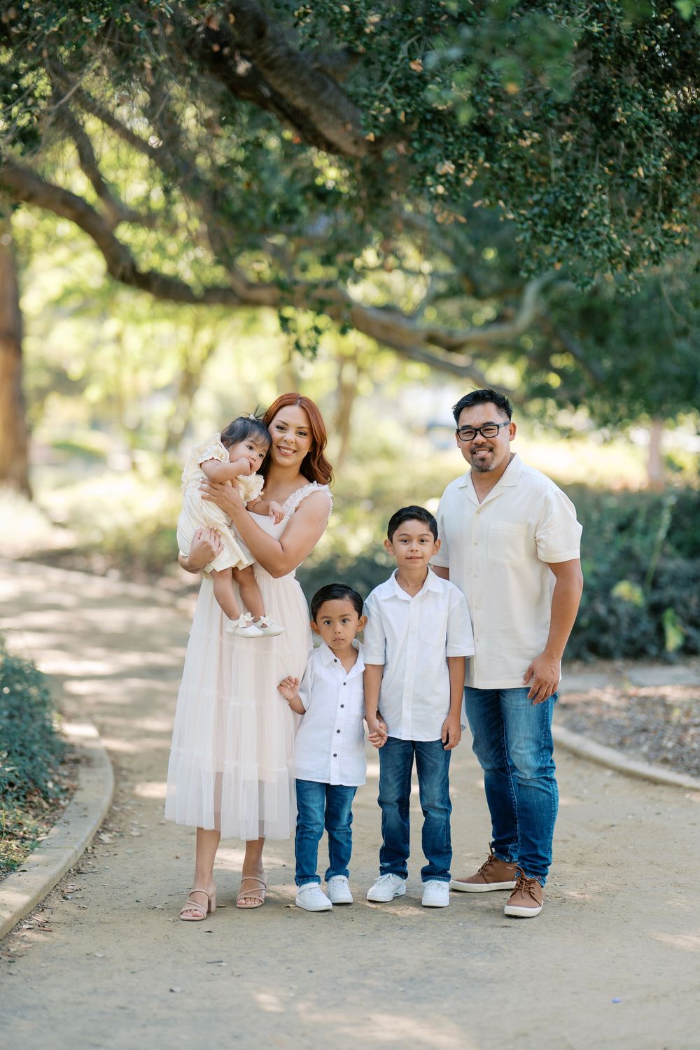 A family wearing coordinated white outfits walks together on a sunlit path in a natural outdoor setting.