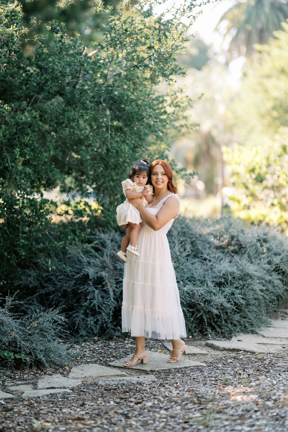 Two people in white dresses embrace while standing on a stone path surrounded by lush greenery in warm sunlight.