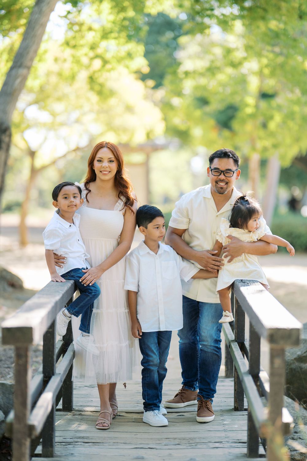 A family in coordinated white and denim outfits poses together on a wooden bridge in a park setting.