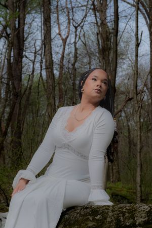 Young woman in white lace dress sits on mossy rock in forest, gazing upward thoughtfully.  Uniontown, PA