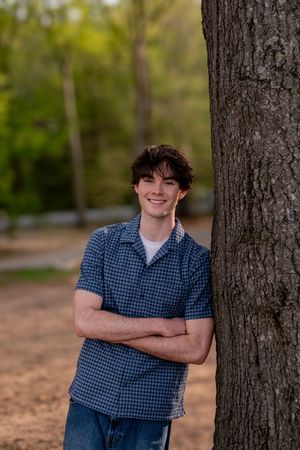 A smiling teenage boy with curly hair leans against a tree with arms crossed, wearing a blue plaid shirt outdoors.