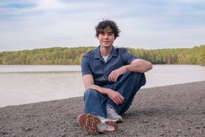 Young man sitting on a rocky shoreline with a calm river and lush green trees in the background.