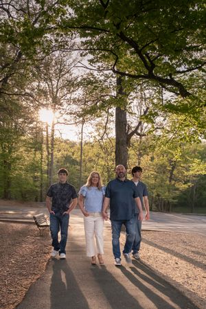 Family of four walking on a sunlit park path surrounded by lush green trees during golden hour.