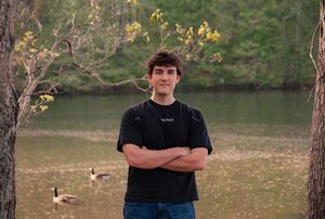 Young man with crossed arms standing by a serene pond with geese, surrounded by trees in autumn.