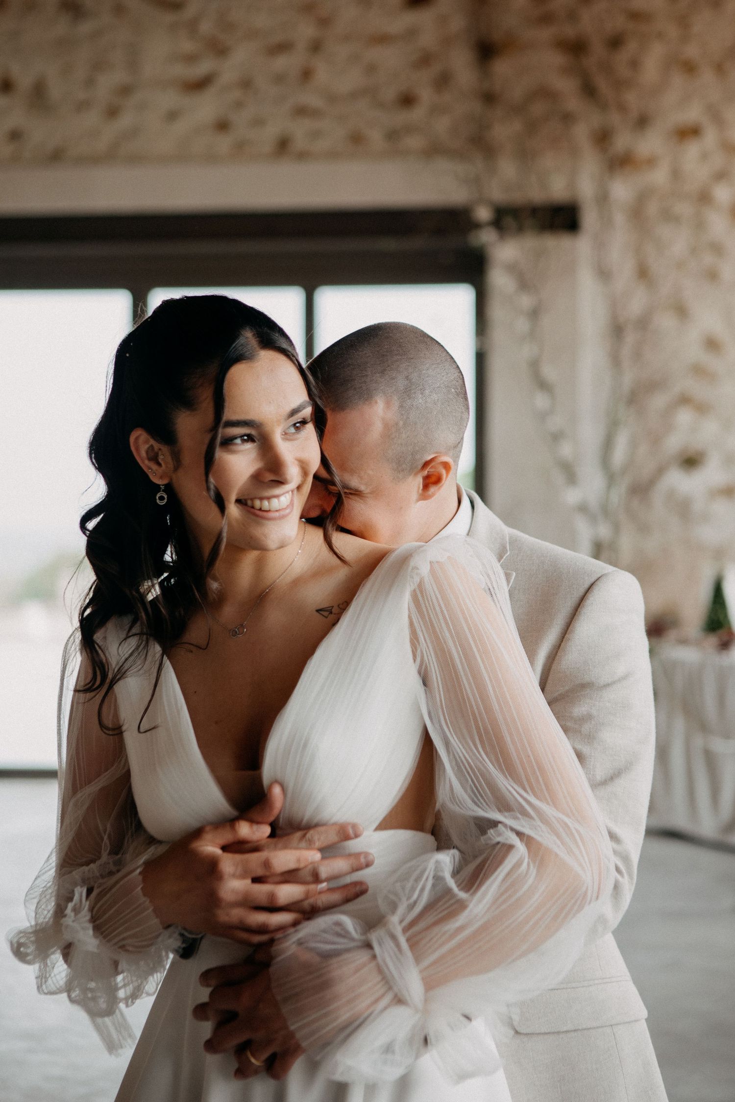 Groom embraces smiling bride in a sheer white gown in a rustic stone-walled wedding venue.
