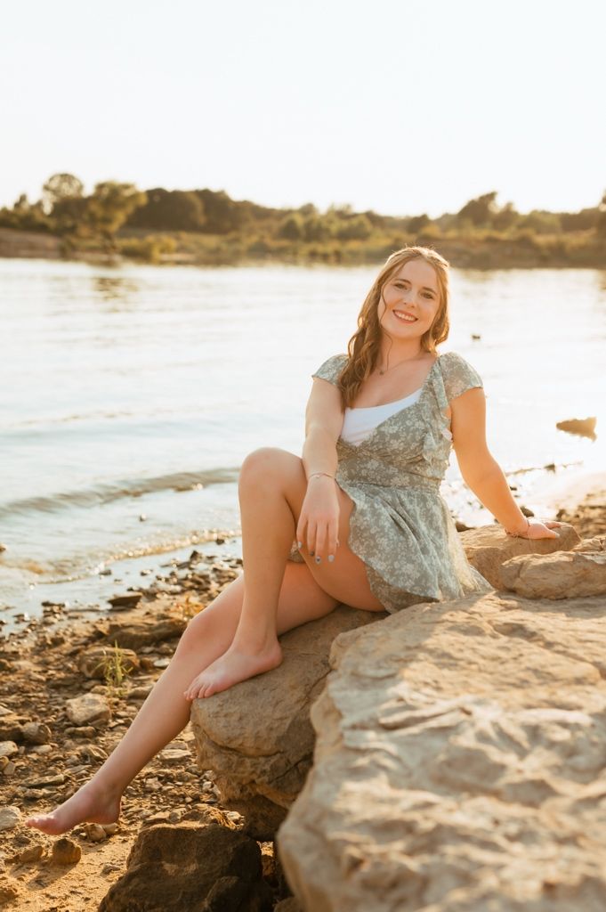 Person sitting barefoot on rocky beach at golden sunset wearing floral dress at shoreline.