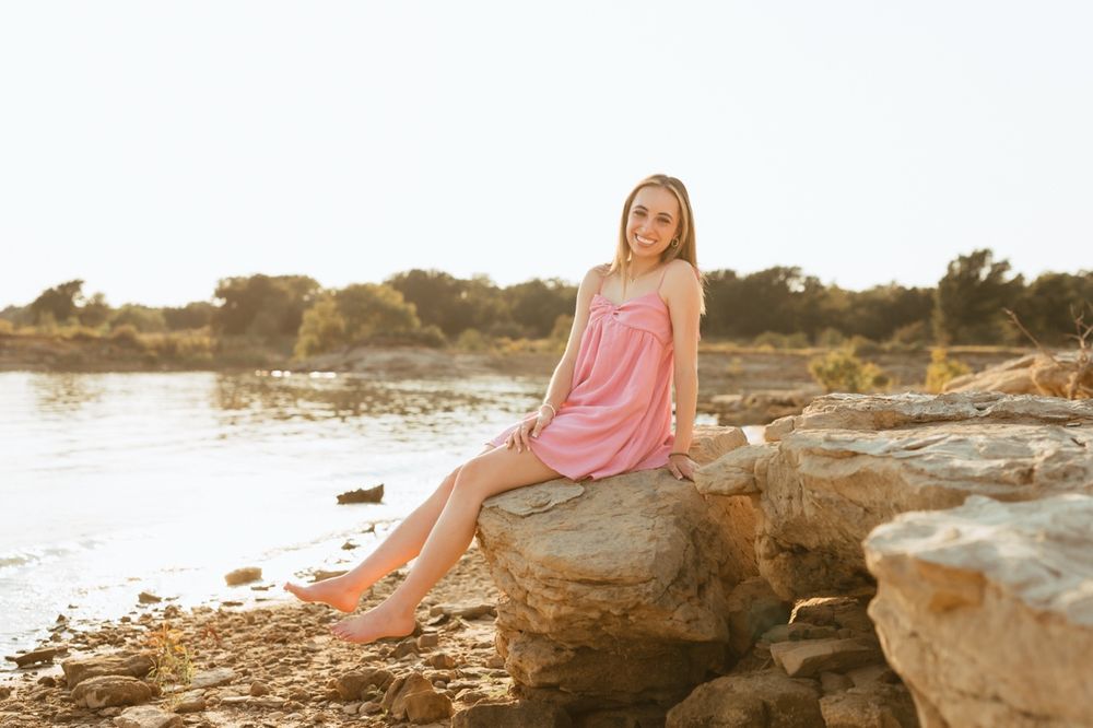 A woman in a pink dress poses outdoors on rocky coastal cliffs at golden hour.