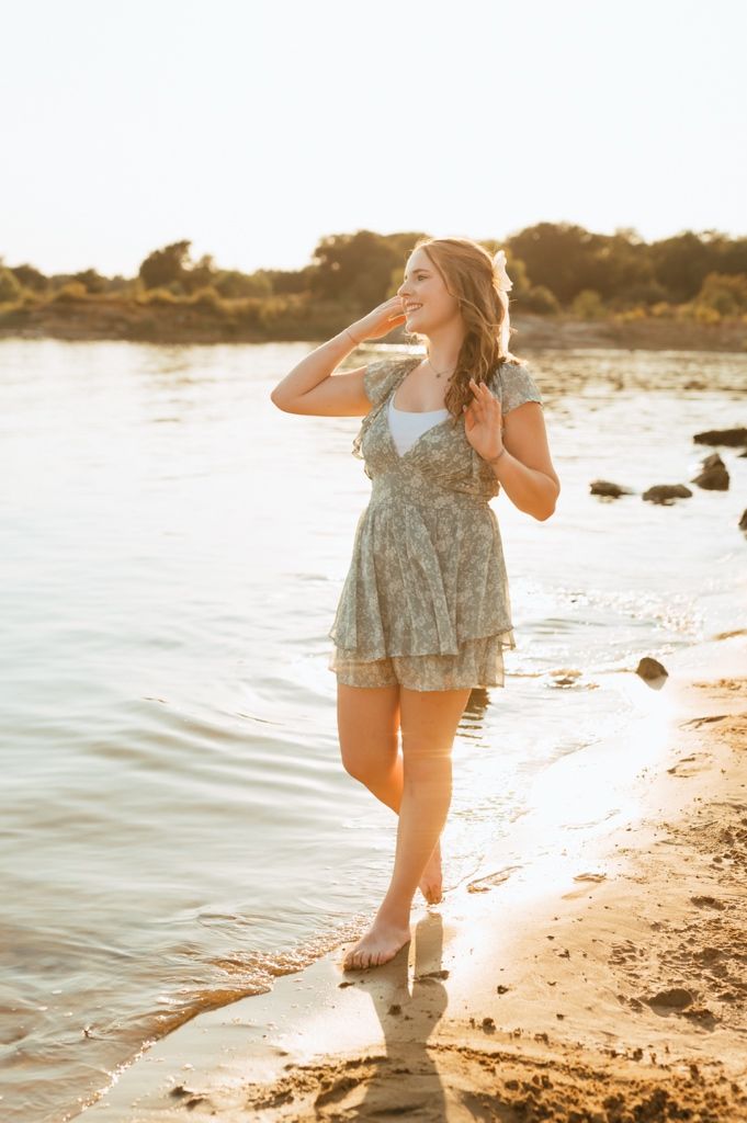 A figure in a flowy dress and boots poses gracefully by the water's edge at golden hour on a rocky beach.