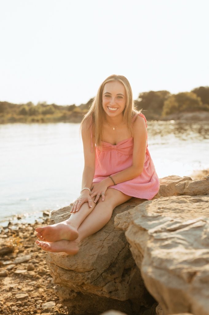 A woman in a pink dress sits on coastal rocks at sunset, creating a dreamy summer beach portrait.