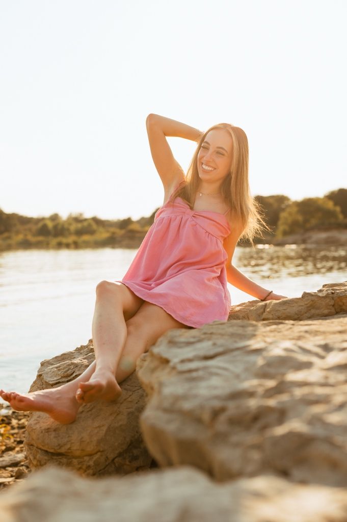 A person in a pink dress poses gracefully on coastal rocks at golden hour as sunlight creates a warm glow.