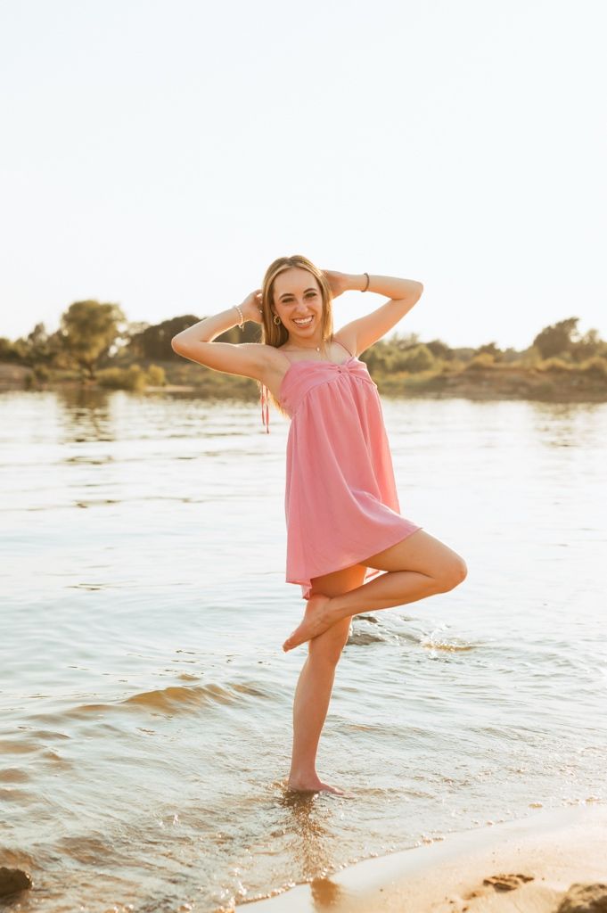 A person in a pink sundress poses playfully at the beach during golden hour, standing in shallow water near the shore.