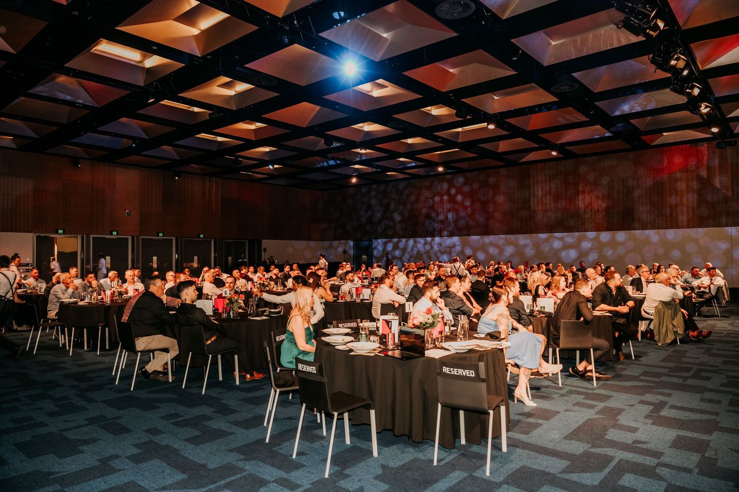 Conference attendees sit in rows of chairs in a large modern event hall with dramatic lighting and exposed ceiling beams.