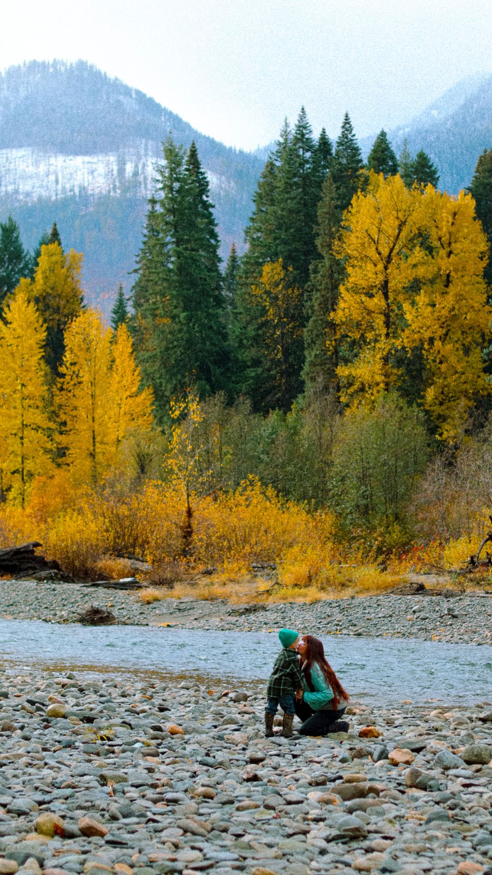 Person crouching on rocky riverbank surrounded by vibrant yellow autumn trees and misty mountains.