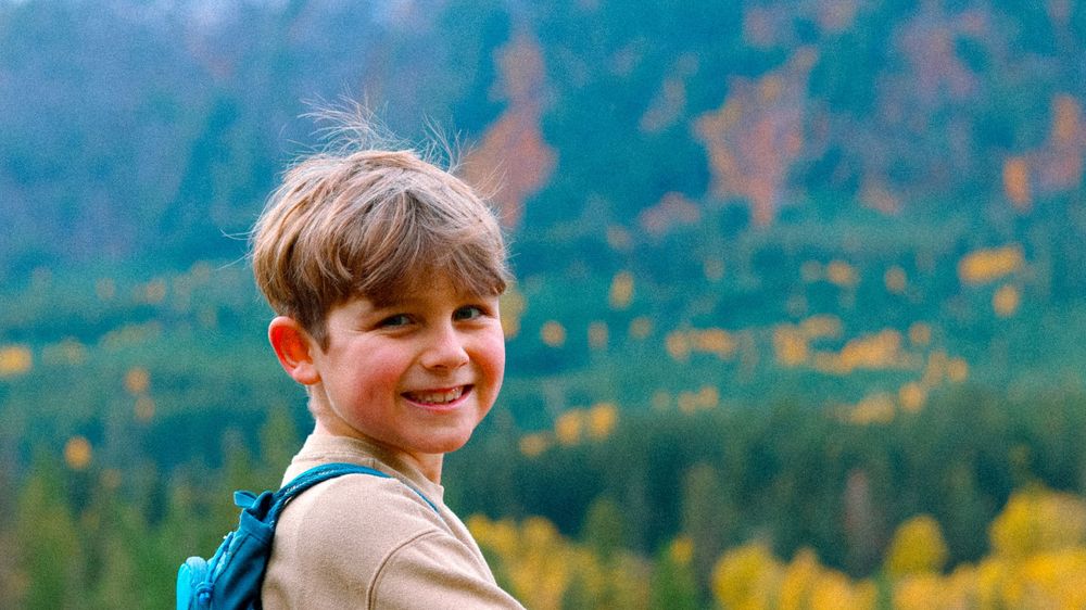 Young boy smiling outdoors with a mountain forest backdrop during autumn season.