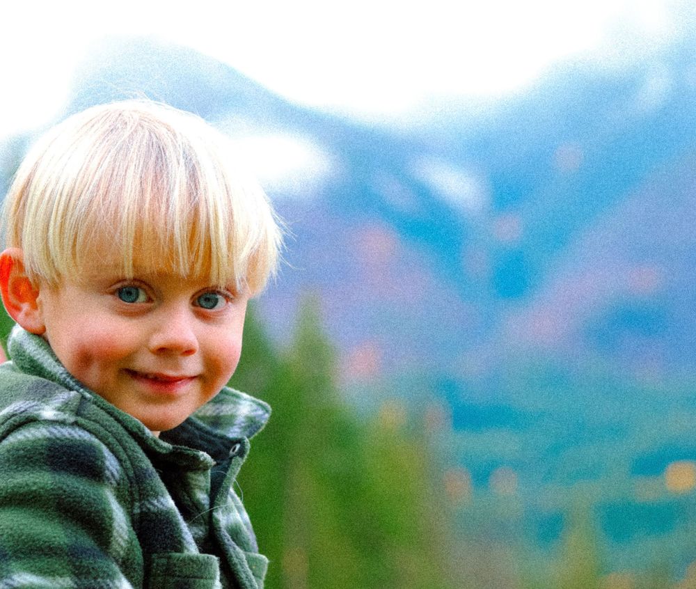 Blond toddler smiling with misty mountain and evergreen trees in soft-focus background.
