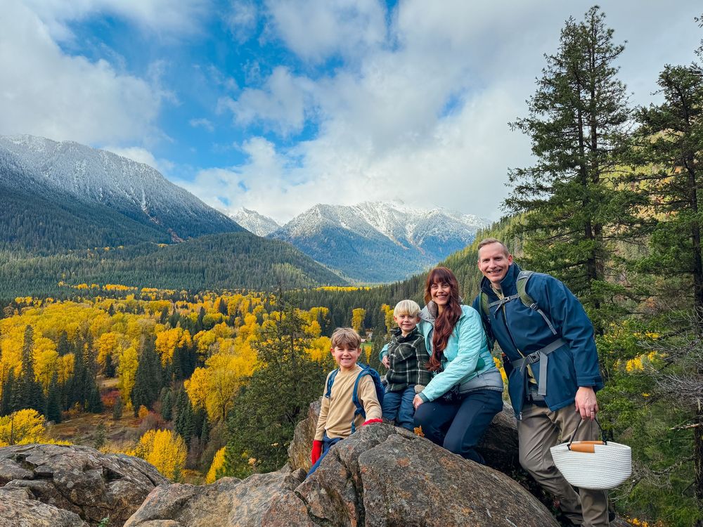 Family of four posing on rocks with stunning snow-capped mountains and fall foliage behind them.
