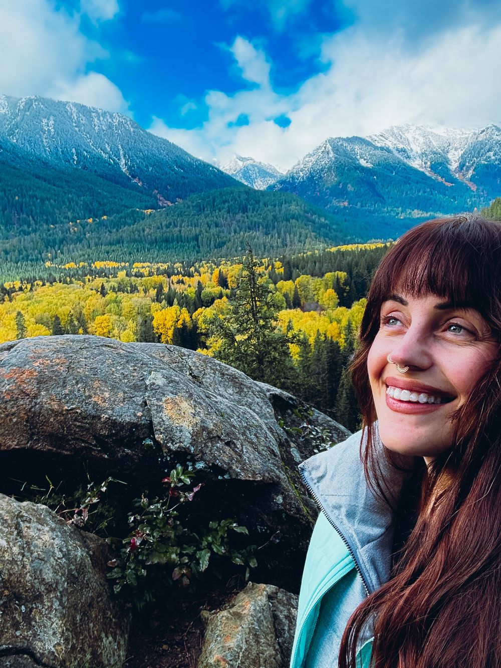 Woman smiling on a rocky overlook with snow-capped mountains and golden autumn trees in background.