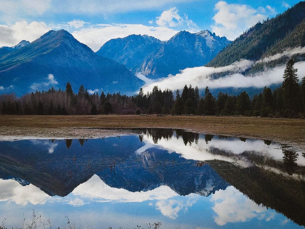 Majestic mountain range reflecting perfectly in a calm alpine lake surrounded by pine trees and misty clouds.