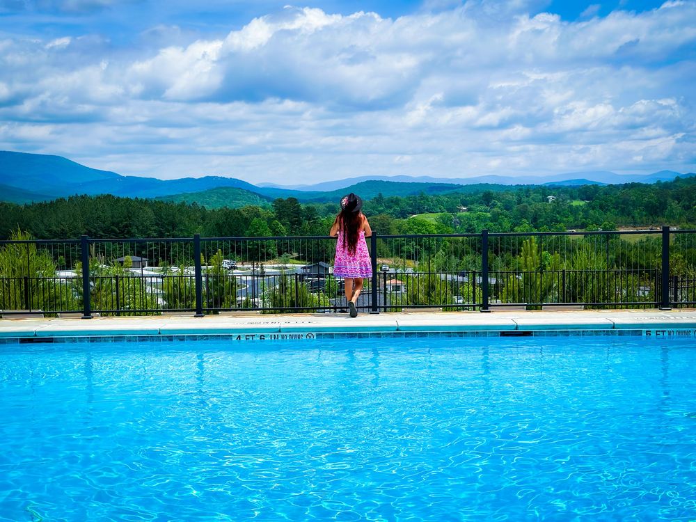 Woman in pink dress standing by infinity pool overlooking scenic green mountain valley under cloudy sky.