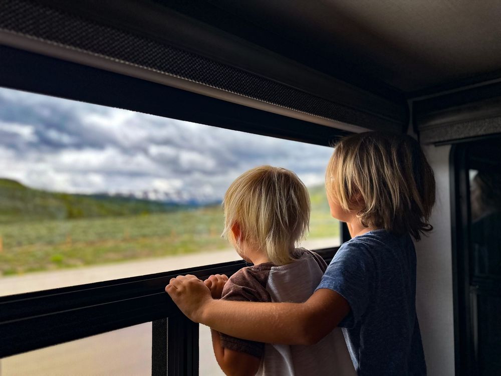 Two young blonde children looking out an RV window at a cloudy mountain landscape together.