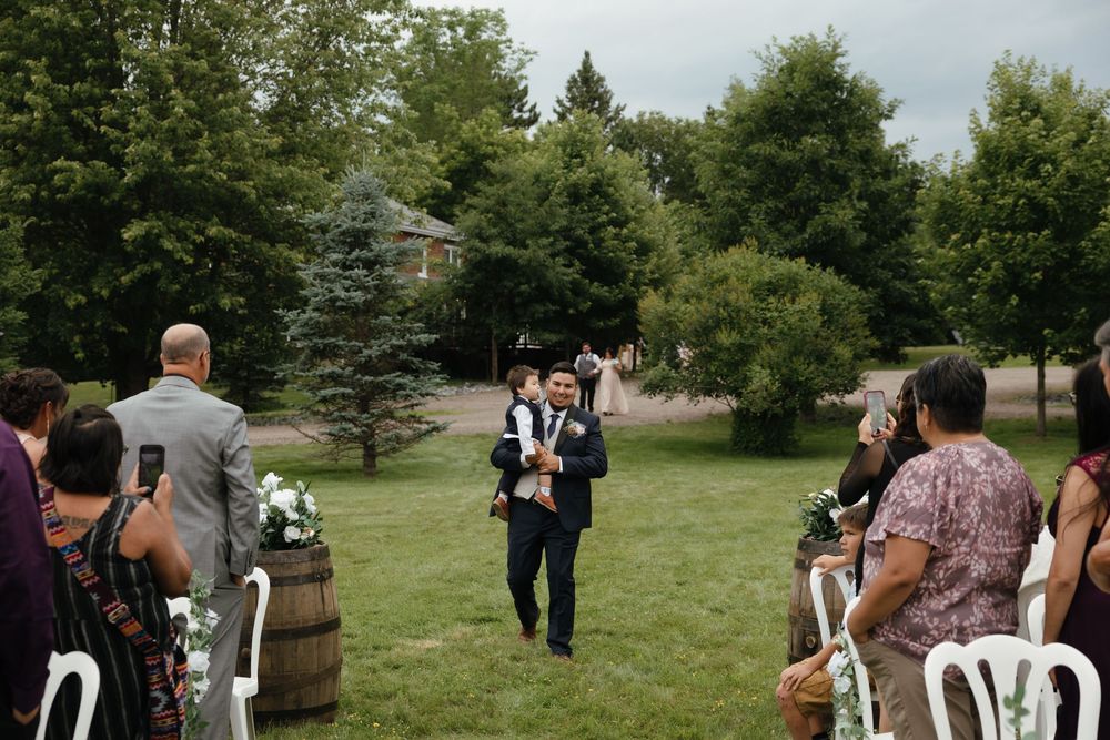 A wedding ceremony takes place outdoors on a beautiful green lawn with guests seated in folding chairs.