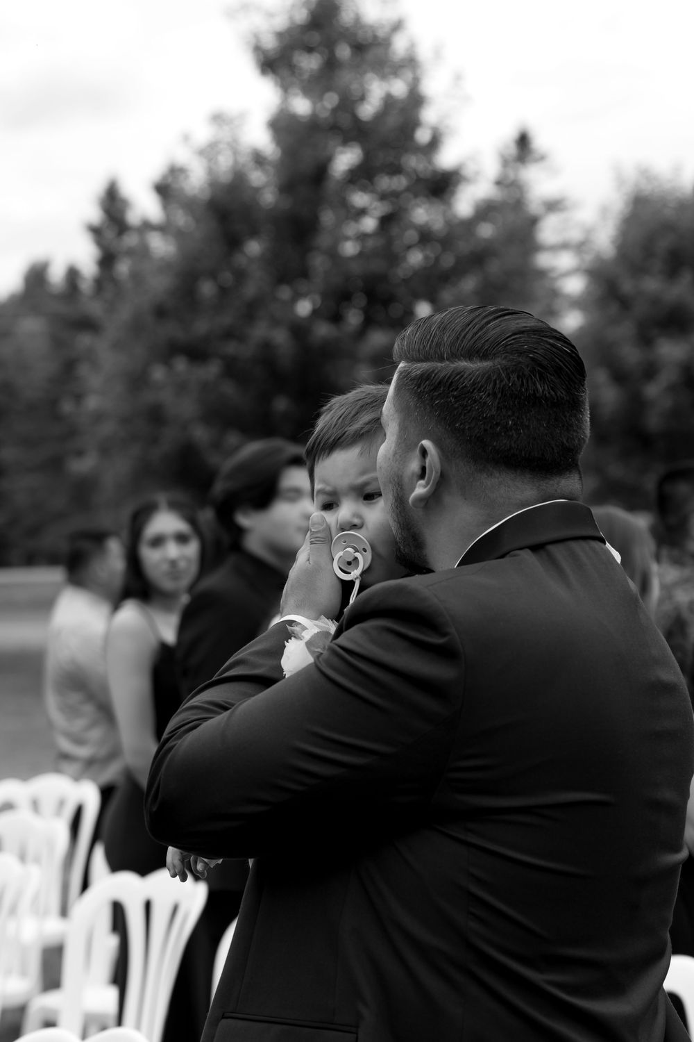 A tender black and white moment at an outdoor wedding ceremony with a baby in formal attire.