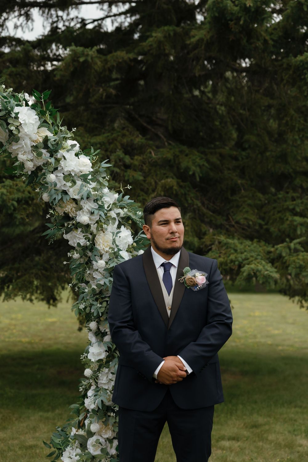 Wedding ceremony setup outdoors with an elegant white floral arch and evergreen trees in the background.