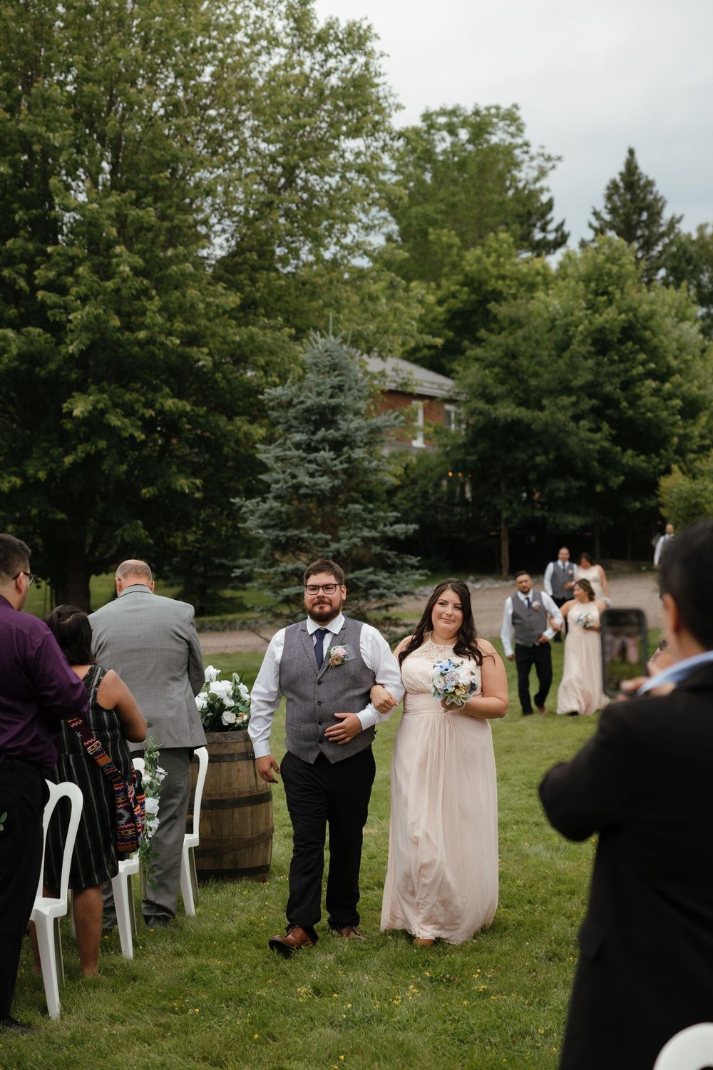 Wedding guests gather outdoors on a lawn with white chairs and evergreen trees in the background.