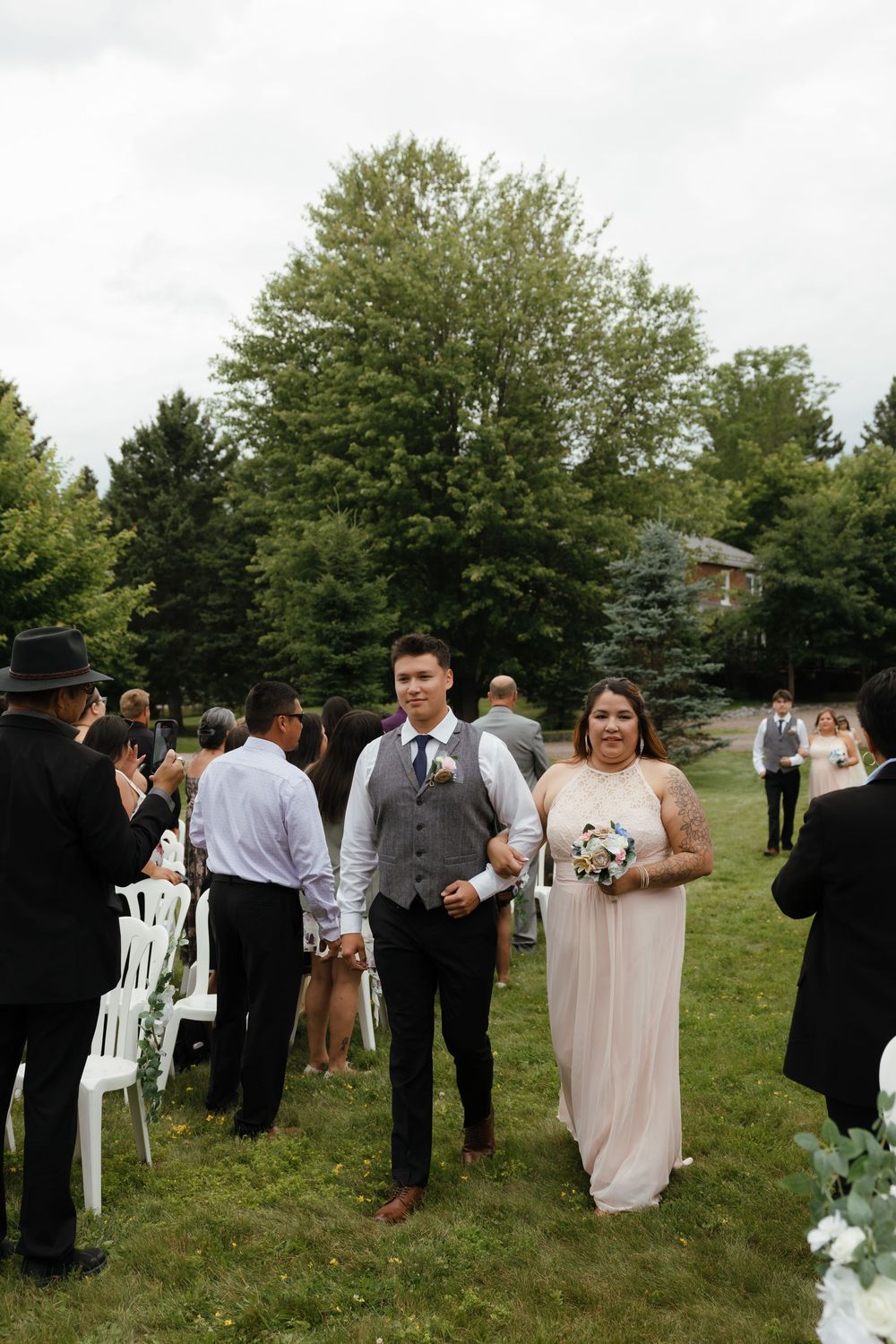 Wedding guests mingle on a green lawn with white chairs during an outdoor reception among tall trees in summer.