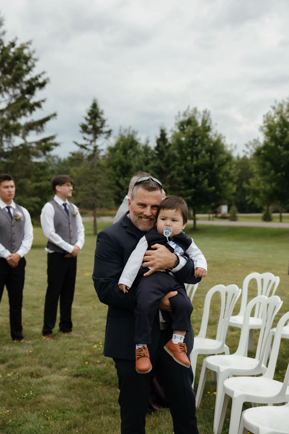 White chairs are set up on a grassy field with groomsmen in dark suits standing nearby.
