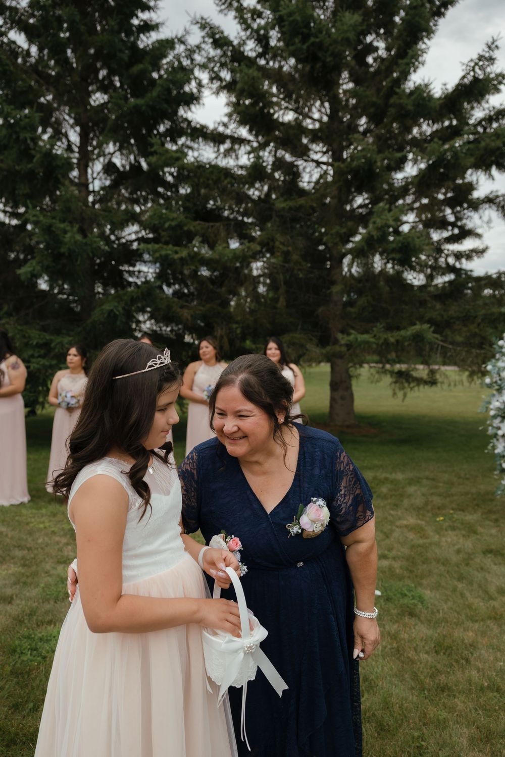 Outdoor wedding scene with attendees in formal attire gathering near evergreen trees and floral decorations.