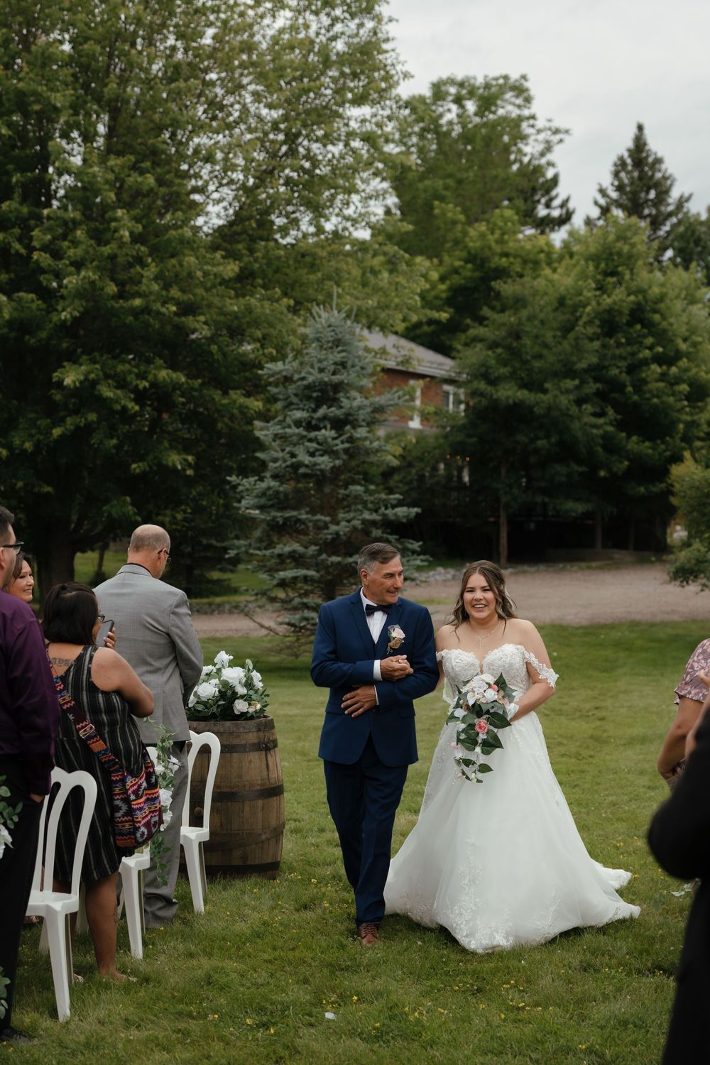 A wedding ceremony takes place outdoors on a grassy lawn with white chairs and rustic barrel decor near evergreen trees.