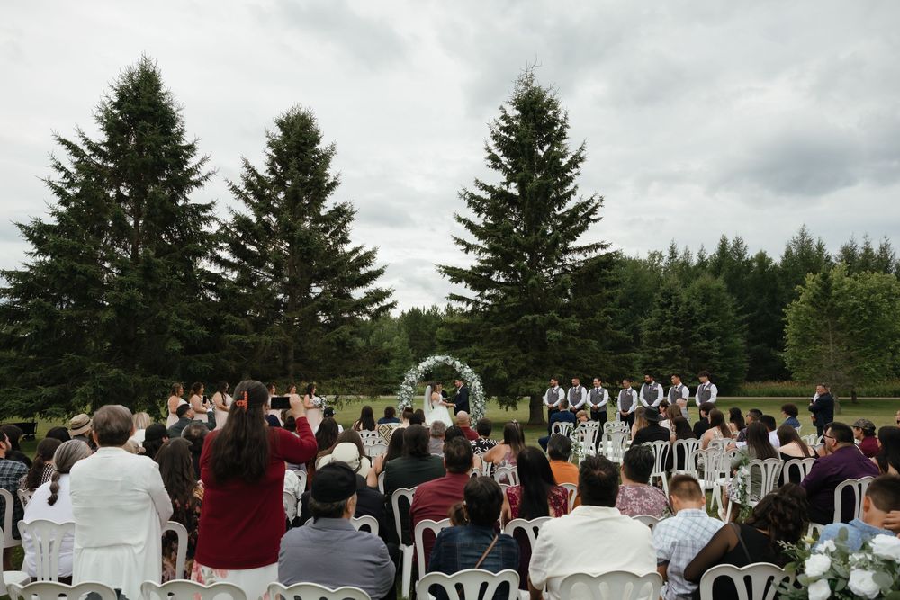 An outdoor wedding ceremony in progress with evergreen trees and cloudy skies as the backdrop.