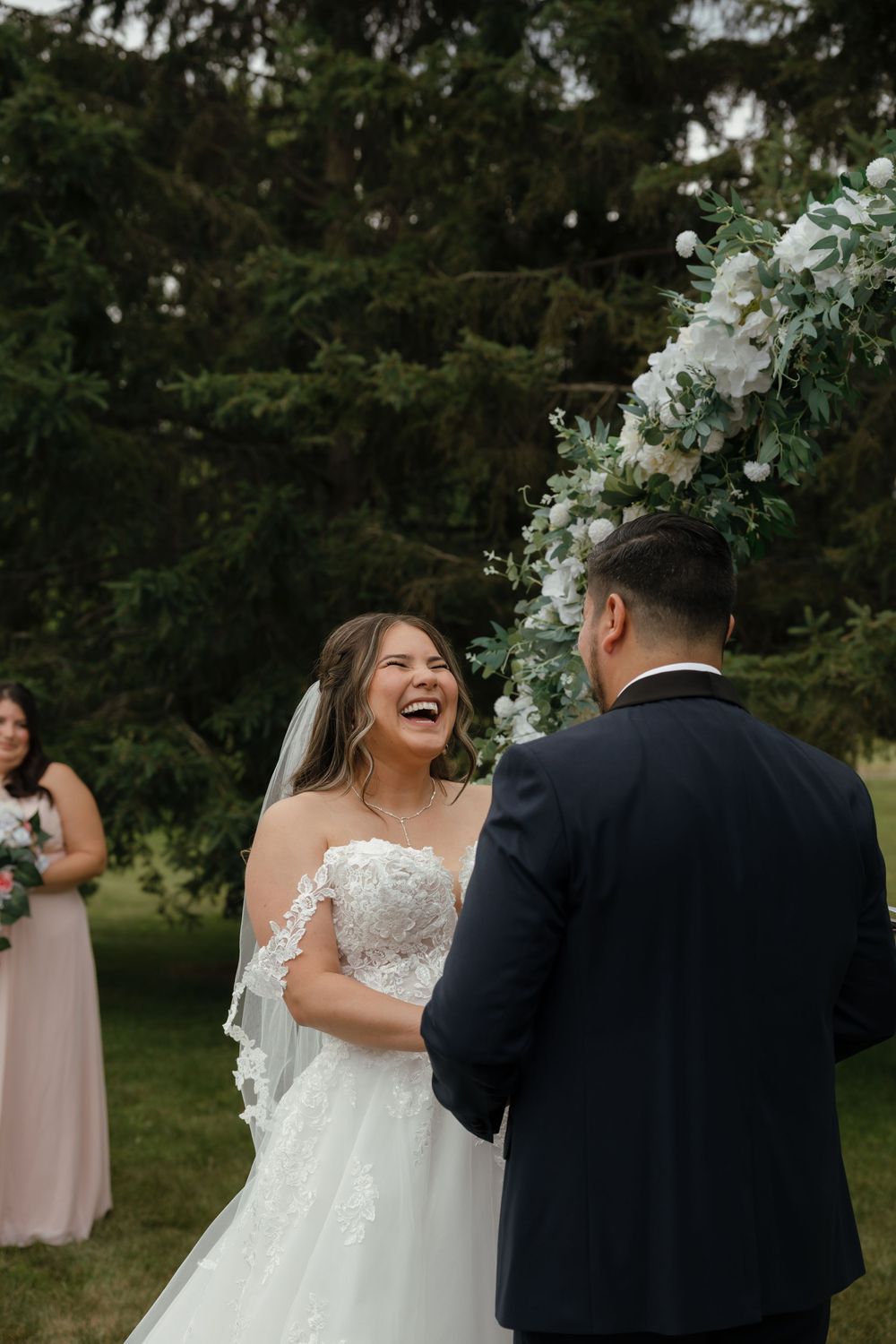 A joyful wedding moment captured outdoors with the bride in white lace and groom in dark suit against evergreen trees.