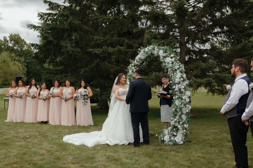 Wedding ceremony panorama shows floral arch and bridal party standing in rows across grassy outdoor venue.