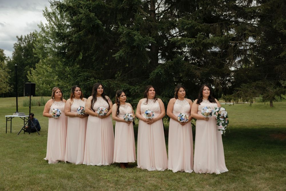 A group of bridesmaids in blush pink dresses pose together in an outdoor garden setting for wedding photos.
