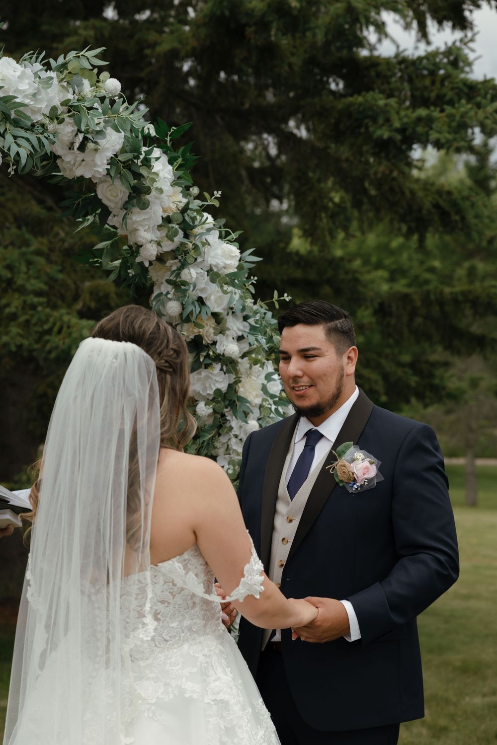 A couple exchanges vows under a beautiful white floral arch during an outdoor wedding ceremony.