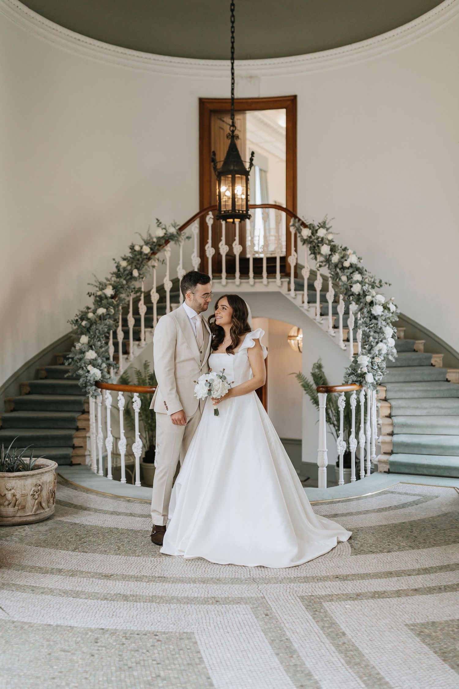 A romantic bridal couple sharing intimate moments on an elegant curved staircase decorated with white floral arrangements.