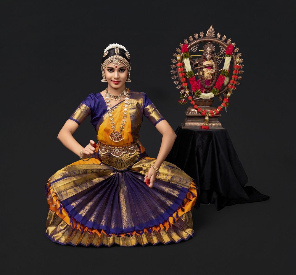 A dancer performs classical Indian Bharatanatyam dance poses in a purple and gold sari against a black background.