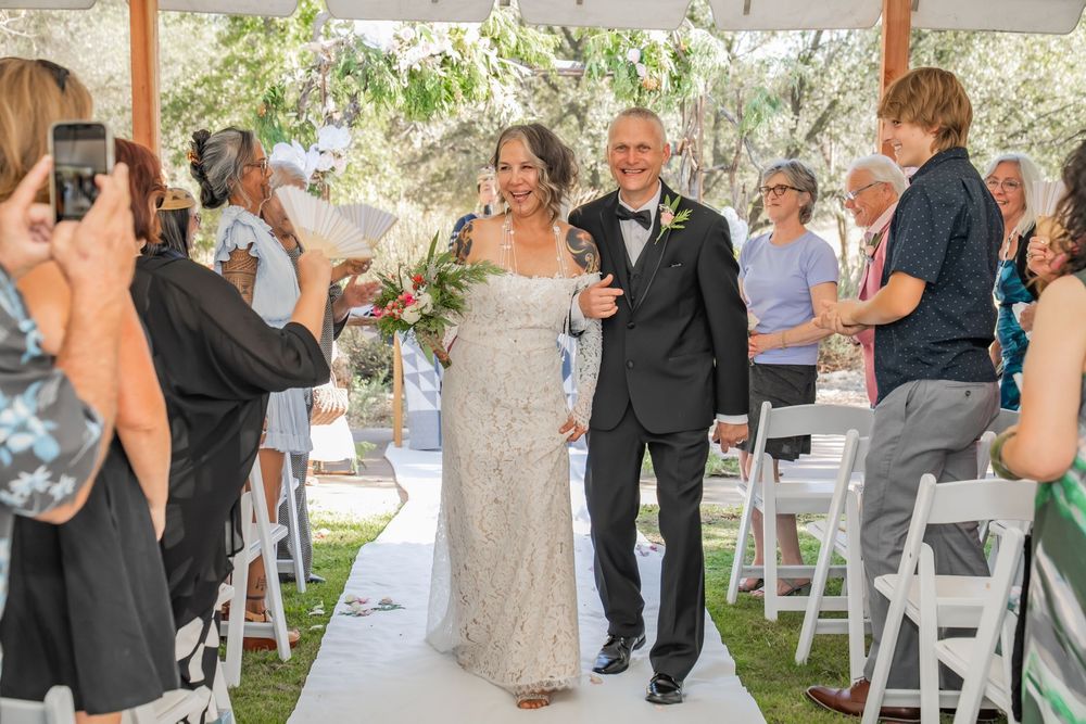 A joyous moment captured at an outdoor wedding ceremony as two people walk down the aisle while guests take photos.