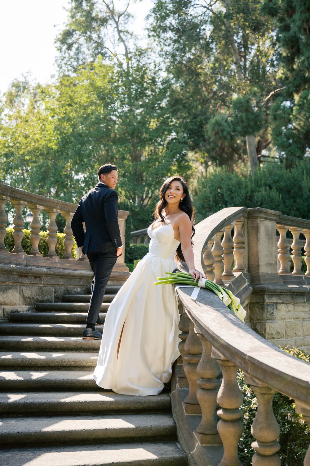 A couple in wedding attire poses along an elegant stone balustrade staircase in a scenic outdoor garden setting.