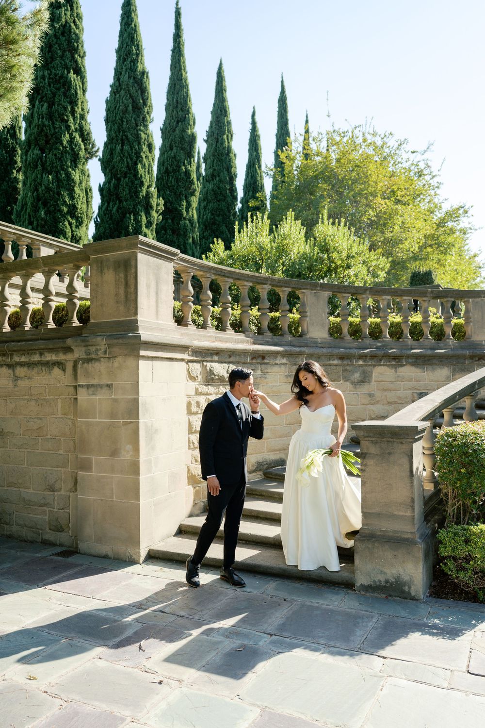 A couple in wedding attire posing in an elegant stone courtyard with cypress trees in the background.