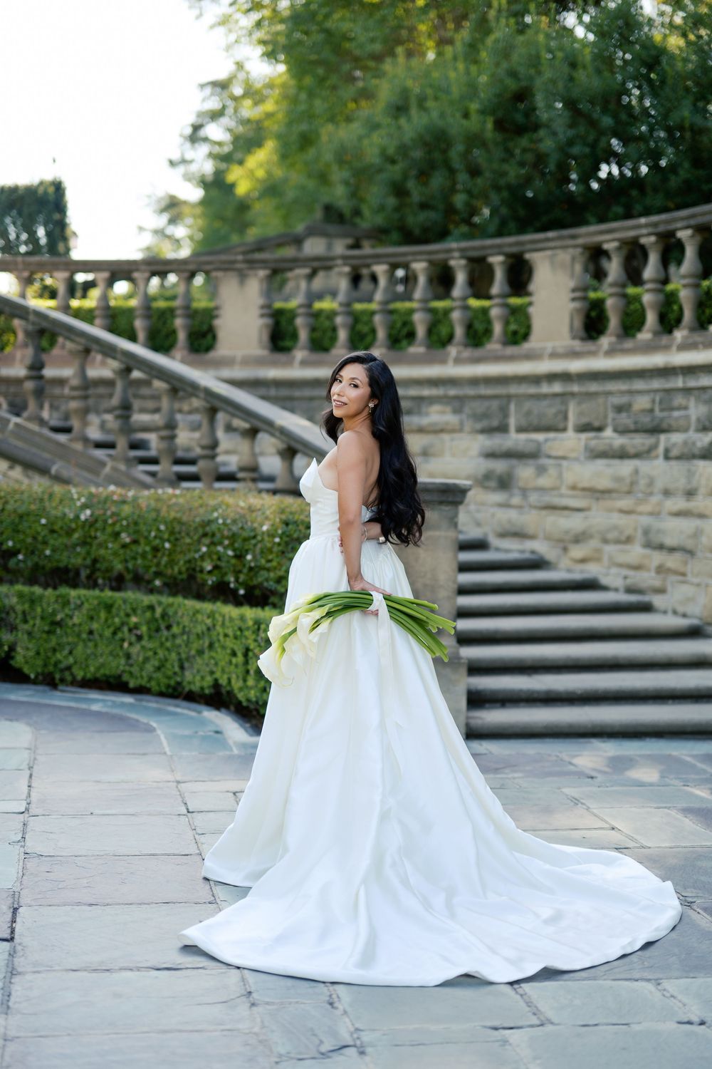 A bride in a flowing white ballgown poses gracefully on stone steps surrounded by Italian cypress trees.