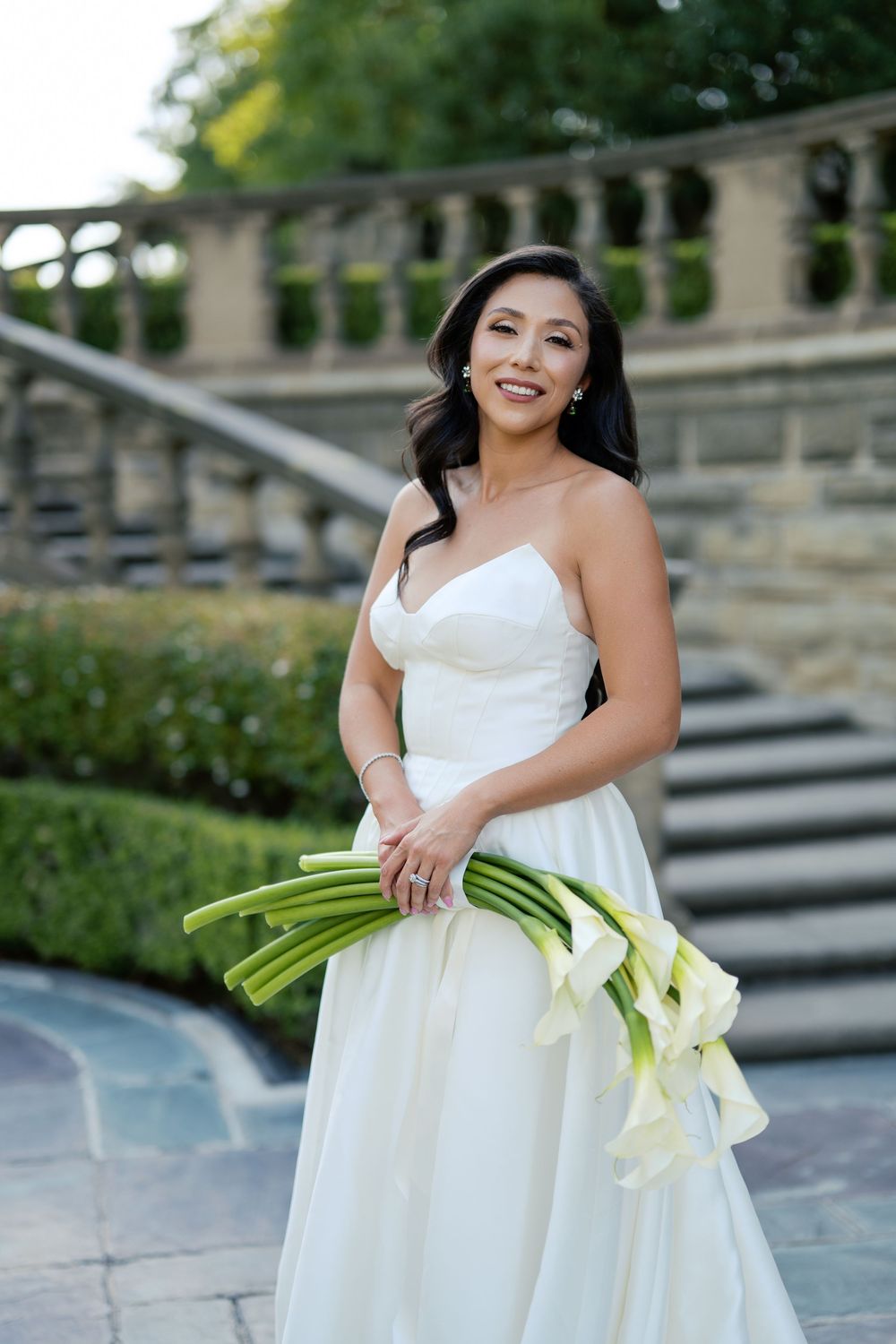A bride in a white strapless wedding gown holds a bouquet of green-stemmed calla lilies on stone steps.
