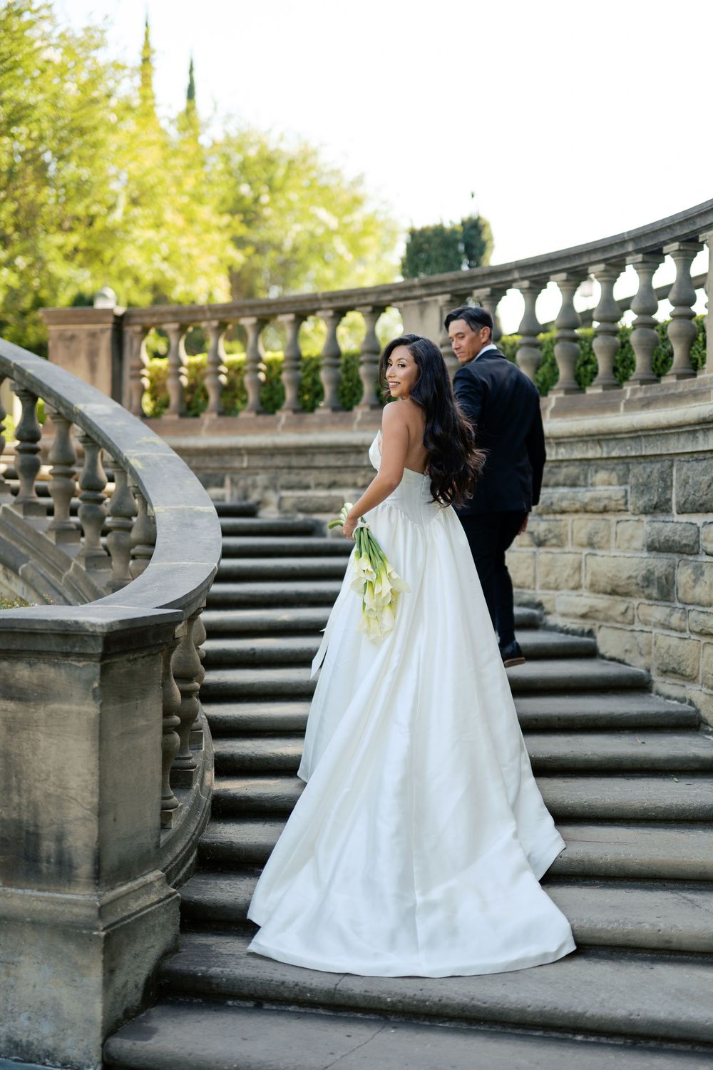 Wedding couple posing on grand stone staircase with ornate balusters in garden setting.