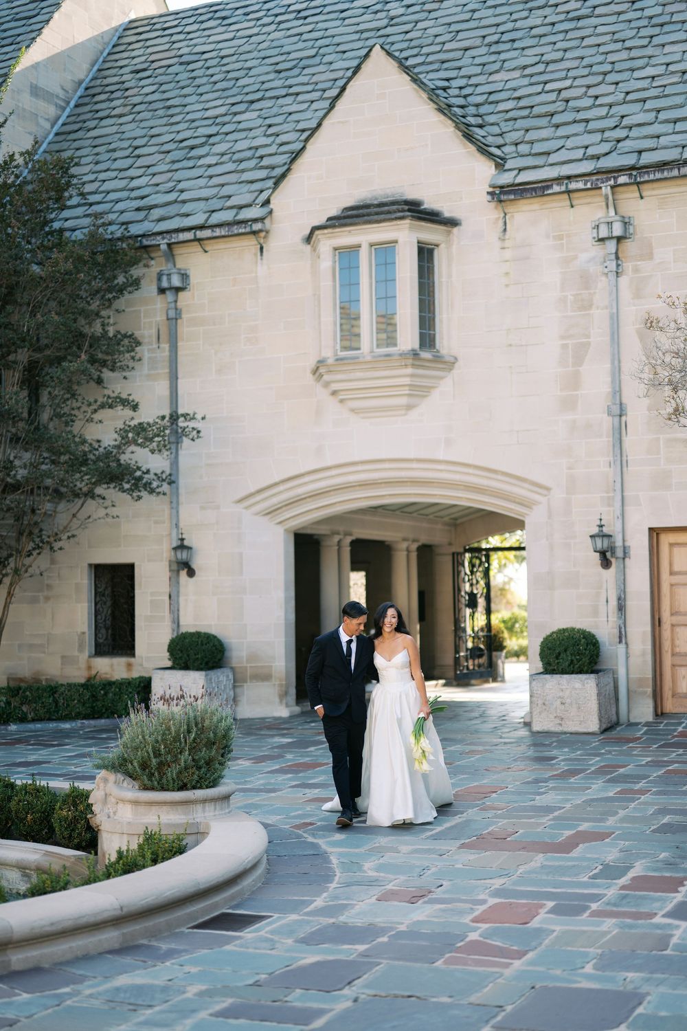 A couple poses in front of a grand white stone mansion with elegant archways and slate roof.